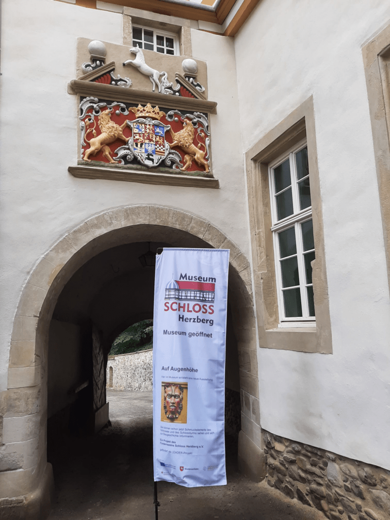 Vertikales Banner vor dem Museum Schloss Herzberg mit Hinweis auf die entstehende Ausstellung „Auf Augenhöhe“, inklusive Logo, Projektbeschreibung und Förderlogos von EU, Niedersachsen und LEADER‑Region.